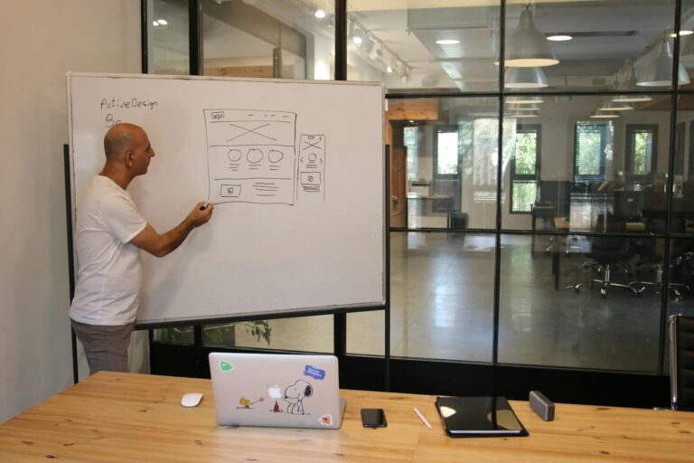 Side view of adult male manager in white t shirt standing near whiteboard and explaining business strategy during work in modern office